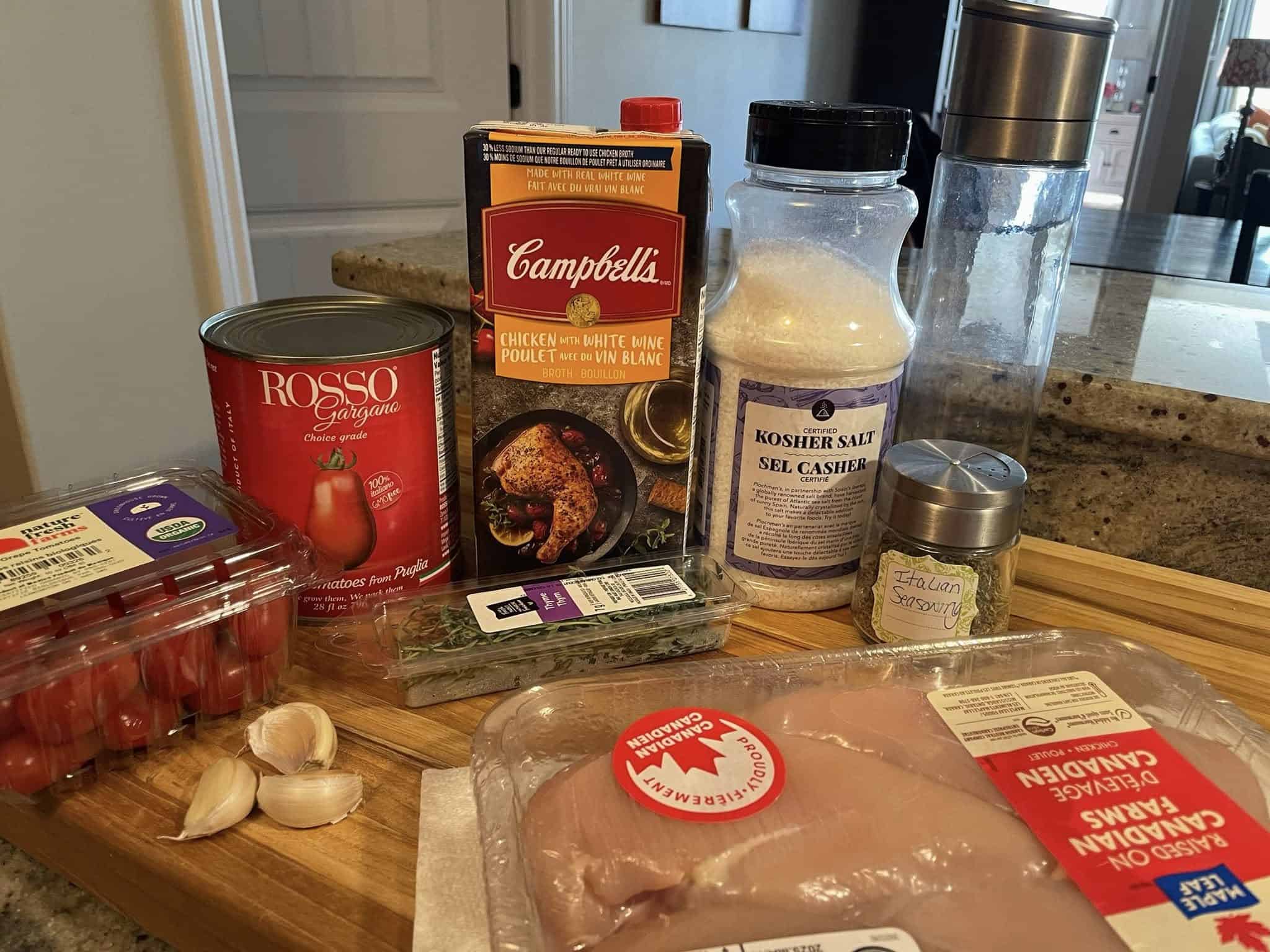 Ingredients laid out for baked chicken pasta with cherry tomatoes, including garlic cloves, cherry tomatoes, canned peeled tomatoes, chicken breasts, chicken broth, kosher salt, Italian seasoning, and fresh thyme.