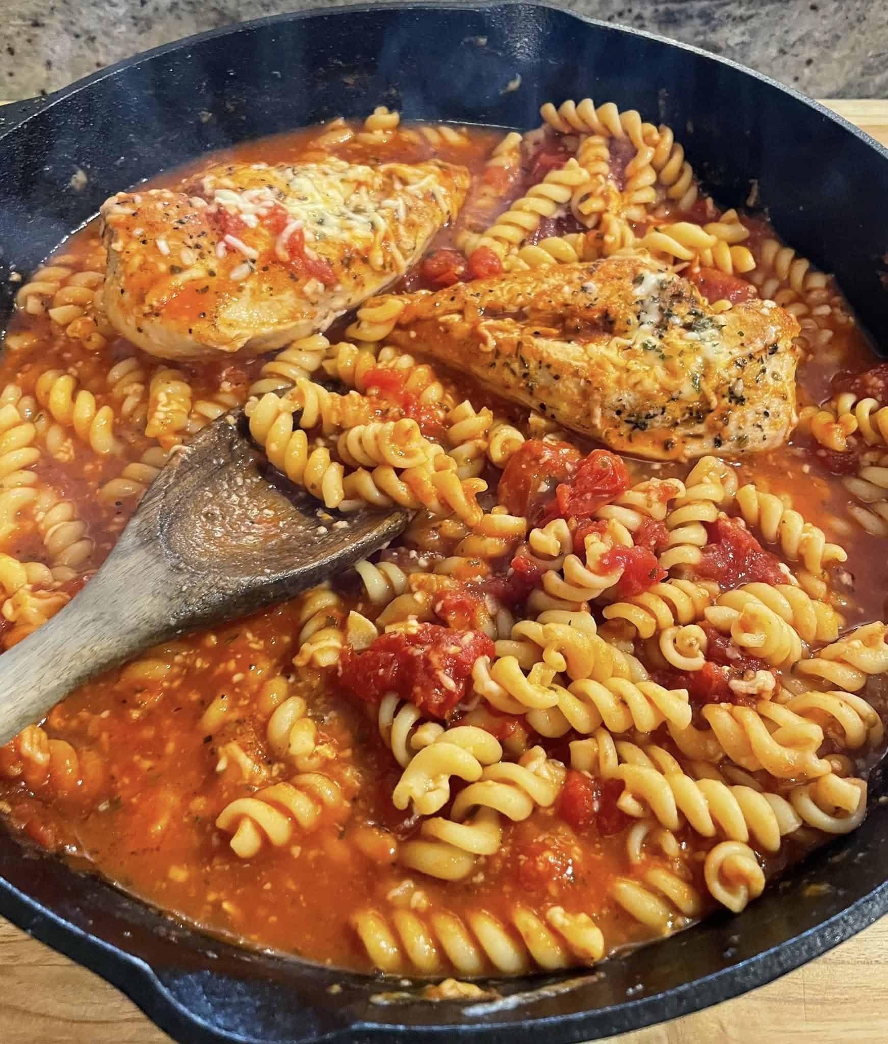 Wooden spoon stirring baked chicken pasta in a skillet with rich tomato sauce